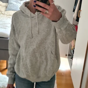 Ljusgrå hoodie med ficka - Säljer en enkel och stilren ljusgrå hoodie med huva och snören. Tröjan har en stor magficka och ribbade muddar vid ärmslut och nederkant. Perfekt för en avslappnad look. Säljs för kmr it till användning💕