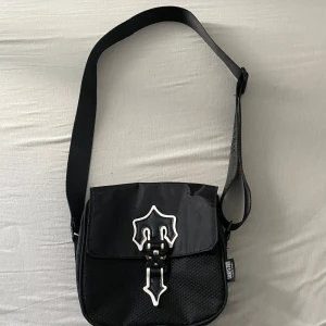 Säljer en svart axel väska - märke trapstar  - SÄLJER: Unik axelväska – streetstyle med edge | 1000 kr  Letar du efter den perfekta väskan som sticker ut utan att skrika? Den här svarta axelväskan med detaljer i silver har exakt rätt blandning av stil, funktion och attityd. Designen är exklusiv, med en unik låsdetalj som påminner om en rustning – för dig som inte kompromissar med din stil. Ej äkta ! Ej använd och man kan skjustera väskans size.