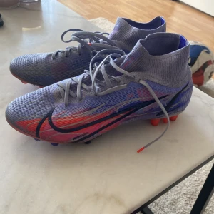 Nike Mercurial superfly 8 elite i MBAPPE versionen - Säljer ett par Nike Mercurial fotbollsskor i lila med röda och svarta detaljer. Skorna har snörning, högre skaft och mönstrad ovandel. Sulan har röda och lila dobbar för bra grepp på planen. Perfekta för dig som vill sticka ut på fotbollsplanen!