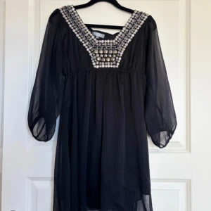 Svart klänning med pärldetaljer - Elegant svart klänning från Christelle med imponerande pärldetaljer runt halsringningen. Klänningen har trekvartsärmar och en luftig passform, perfekt för en stilfull look.