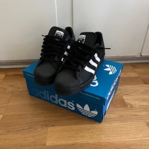 Svarta Adidas Superstar 82 - Klassiska svarta Adidas Superstar 82 sneakers med vita ränder och svart sula. 