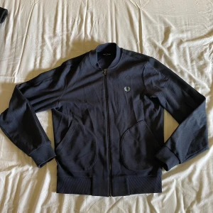 Mörkblå track jacket från Fred Perry - Säljer en marinblå track jacket från Fred Perry med dragkedja och ribbade muddar. Loggan är ljublå och längs armarna syns ljusblåa detaljer i vissa vinklar. Denna tröjan är fan assnygg och i bra condition.