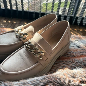 Beige loafers med kedjedetalj - Snygga beige loafers med en iögonfallande guldkedja över vristen. Perfekta för att ge en stilren touch till din outfit.