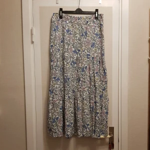 Blommig långkjol från C&A strl XS/ S - Charmig långkjol från C&A med ett vackert blommönster i blått, gult och rosa på en vit bakgrund. Kjolen har en bekväm elastisk midja och är perfekt för en somrig look. Den luftiga designen gör den idealisk för varma dagar.