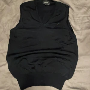 Svart väst från Boomerang - Säljer en stilren svart väst från Boomerang med v-ringning. Perfekt för att lägga till en extra touch till din outfit. Västen har en liten broderad logga på bröstet. Den här därimot ett litet hål vänster hörn nere men det går att laga. Den är i 100% Mariano ull 