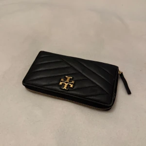  plånbok från Tory Burch - Snygg svart quiltad plånbok från Tory Burch med dragkedja och ett elegant guldemblem på framsidan. Perfekt för att hålla ordning på dina kort och kontanter med stil.