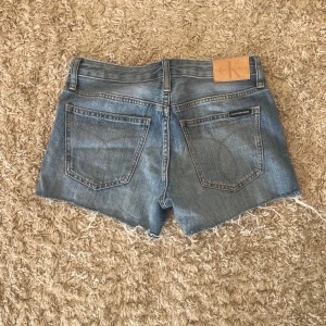 Blå jeansshorts från Calvin Klein - Säljer ett par klassiska blå jeansshorts från Calvin Klein med råa, fransiga benslut och femficksdesign. Shortsen är i mid waist, perfekt för sommardagar. Ikonisk Calvin Klein-lapp bak i midjan.