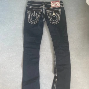 Mörkblå True Religion jeans med vita sömmar - Säljer ett par mörkblå True Religion jeans med tydliga vita kontrastsömmar och klassiska bakfickor med broderad logga. Jeansen är lågmidjade och har lite utsvängning.  Storlek 24 och midja 35cm passar mig perfekt som har 32-34 vanligtvist i jeans.