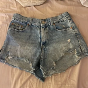 Blå jeansshorts från ZARA - Säljer ett par coola blå jeansshorts från ZARA. De har en avslappnad look med slitna detaljer och fransar vid kanterna. Perfekta för en avslappnad stil i sommar. Storlek 11-12 år.