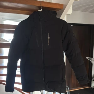 Svart dunjacka från Sail Racing - Säljer en svart dunjacka från Sail Racing med dragkedja och huva. Jackan är tillverkad med Allied Feather & Down, vilket ger bra isolering. Perfekt för kalla dagar. Kan användas av både tjejer och killar. Priset kan givetvis diskuteras. Köpare betalar för frakt. Köp för 2000kr