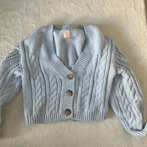 Ljusblå stickad kofta med knappar - Säljer en mysig ljusblå stickad kofta med flätstickat mönster och tre stora knappar framtill. Perfekt för kyliga dagar och ger en stilren look. Passar bra till både jeans och kjol.