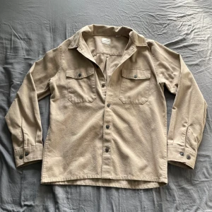 Beige overshirt från Jack & Jones - Säljer en beige overshirt från Jack & Jones med klassisk krage och knäppning framtill. Skjortan har två bröstfickor med lock och knappar samt långa ärmar med manschett. Perfekt att bära som lager över en t-shirt.