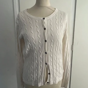 Vit kabelstickad kofta från Park Lane - Säljer en klassisk vit kabelstickad kofta från Park Lane med rund halsringning och knappar framtill. Koftan har långa ärmar och diskret broderad logga på bröstet. Perfekt för lager-på-lager och enkel att matcha med olika outfits.💕