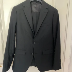 Svart kostym från Riley (kavaj + byxor) med vit skjorta - Säljer en klassisk svart kostym från Riley i slim fit, med matchande vit skjorta och byxor. Kavajen har två knappar, fickor med lock och smal passform. Perfekt för bal eller fest. Skjortan är långärmad och har klassisk krage. Kavajen är i storlek 46. Byyxor är storlek 44. Skjortan är storlek small.