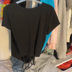 Svart kortärmad topp från Gina Tricot - Snygg svart topp från Gina Tricot med korta ärmar och en trendig snörning baktill.Perfekt för att ge en stilren look med en twist. Tillverkad i mjukt material för extra komfort. Nästan aldrig använd pågrund av fel i storlek.