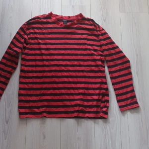 Röd och svart randig tröja från Lindex - Säljer en röd och svart randig tröja från Lindex. Tröjan har långa ärmar och en klassisk rund halsringning. Det är dåck ett litet hål i ärmen men igent märkvärdigt. Perfekt för en avslappnad stil. Hör av dig vid intresse!