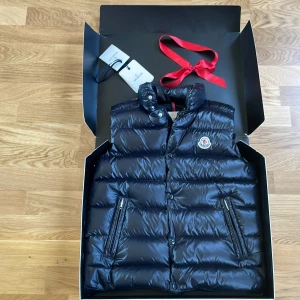 Svart dunväst från Moncler - Säljer en snygg svart dunväst från Moncler. Västen har en quiltad design med dragkedja och knappar framtill. Perfekt för kyligare dagar och kommer i en elegant förpackning från Nordiska Kompaniet.