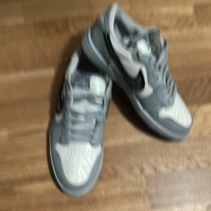 Nike Air Jordans i grått och vitt - Säljer ett par Nike Air Jordans i grått och vitt med svart swoosh-detalj på sidan. Skorna har snörning och rund tå, samt klassisk Air Jordan-logga på hälen. Perfekta för dig som gillar stilrena sneakers med en twist.