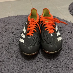 Adidas Predator fotbollsskor med orange detaljer - Säljer ett par Adidas Predator fotbollsskor i svart med vita ränder och orange snörning samt detaljer. Skorna har dobbar för spel på gräs och en sportig design med låg profil. Perfekta för dig som vill sticka ut på planen! Modellen är PRO och är i helt ok skick. Storlek 42 2/3