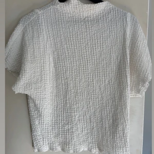 Vit kortärmad blus med struktur och figur sydd med höghals - Säljer en vit blus med korta ärmar och strukturerat tyg. Den är tajt i midjan med lose fit i överdelen. Den har även en höghals. 