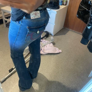 True Religion blå bootcut jeans - Säljer ett par blå jeans från True Religion med bootcut passform och klassiska fickor med rosa broderade detaljer och knapp. Jeansen har en snygg mörkblå tvätt och ikonisk logga på bakfickan. Perfekt för dig som gillar statement-jeans med coola detaljer. Svårt att få tag i helt nya inga skador ❤️