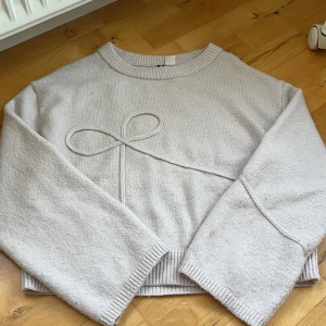 Rosa stickad tröja med rosettdetalj - Säljer en rosa stickad tröja med breda ärmar och en snygg rosettdetalj framtill. Tröjan har ribbade muddar vid hals, ärmslut och nederkant. Perfekt för dig som gillar stilrena och gulliga detaljer.