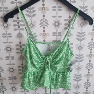 Grön blommig linnetopp med snörning - Söt grön linnetopp med vita små blommor och smala axelband. Toppen har en snygg snörning framtill som ger en cool rynkad effekt och en lätt volang nedtill. Perfekt för dig som gillar färg och mönster!