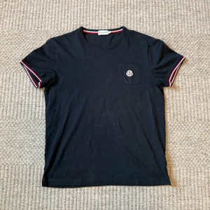 Svart Moncler t-shirt med ficka - Svart t-shirt från Moncler i mjuk bomull. Klassisk rund hals och korta ärmar med snygga röd, vit och blå ränder på mudden. Bröstficka med Moncler-logga ger en clean och exklusiv vibe. Perfekt för dig som gillar stilrena märkesplagg.