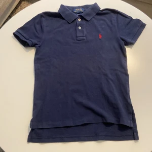Marinblå Polo Ralph Lauren piké - Snygg marinblå piké från Polo Ralph Lauren med klassisk pikékrage och två knappar framtill. Ikonisk röd logga broderad på bröstet. Modellen är kortärmad och har en normal passform. Tillverkad i mjuk bomull som känns skön mot huden.