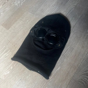 Svart balaclava med goggles från C.P. Company - Säljer en svart balaclava från C.P. Company med inbyggda goggles. Stickad modell i mjukt material som täcker hela huvudet och har öppning för munnen. Perfekt för kalla dagar och streetwear-stil. Ikoniska glasögon framtill ger en unik look. Den har ett litet hål bredvid ögat som man inte ens ser om man har glasen ner