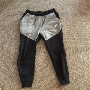 Nike mjukisbyxor svart/grå M - Nike mjukisbyxor i svart och ljusgrått med vit logga på vänster lår. Byxorna har snörning i midjan, ribbade muddar vid benslut och sidofickor. Skön passform och sportig look, perfekta för chill eller träning.