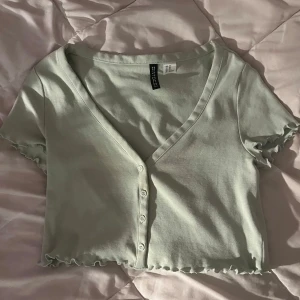 Ljusgrön croppad topp från H&M Divided - Supersöt ljusgrön croppad topp från H&M Divided med v-ringning och korta ärmar. Toppen har små knappar framtill och volangkant vid ärmslut och nederkant.