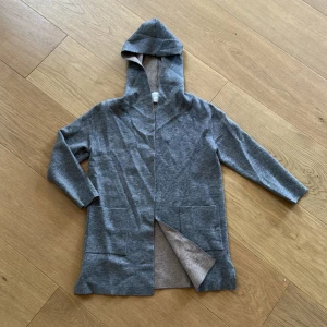 Grå cardigan jacka med huva från Zara - Snygg grå cardigan jacka från Zara med huva och långa ärmar. Jackan har två stora fickor framtill och slitsar i sidorna för extra rörelsefrihet. Mjuk och skön i bomullsmaterial, perfekt för chill dagar. Enkel och stilren design som funkar till det mesta.