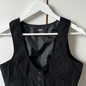 Svart väst från H&M med knappar - Svart väst från H&M i storlek 170. Västen har en klassisk v-ringad design med tre knappar framtill och två dekorativa fickor. Materialet är bomull och den har en stilren, enkel look som funkar till många outfits.