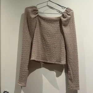 Croppad topp - Säljer en beige croppad topp från H&M med långärmad design och snyggt rynkade axlar. Materialet är mjukt och har en ribbad struktur.