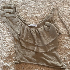 Beige/grön volang topp från Zara - Säljer en beige/grön topp från Zara med volangdetaljer och en axelbandslös design. Toppen har en asymmetrisk form med en axel och är tillverkad i ett mjukt, strukturerat bomullsmaterial. Perfekt för dig som gillar trendiga och unika plagg. Oanvänd.