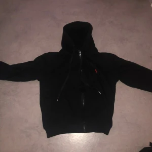 Svart hoodie med kedja från Polo Ralph Lauren - Säljer en svart hoodie från Polo Ralph Lauren i storlek M. Tröjan har dragkedja framtill, huva med snören och klassisk passform. Perfekt för en avslappnad stil och enkel att matcha med det mesta.