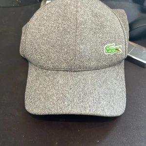 Grå keps från Lacoste - Snygg grå keps från Lacoste med klassisk krokodillogga i grönt och rött framtill. Kepsen har böjd skärm och justerbar rem baktill för perfekt passform. Materialet är mjukt och känns som bomull med en lätt struktur. Perfekt för dig som gillar stilrena accessoarer.