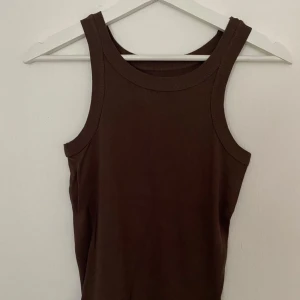 Brun ribbad linnetopp från Lager 157 - Brun ribbad linnetopp från Lager 157 i storlek S. Toppen är ärmlös med breda axelband och har en rundad halsringning. Tillverkad i 100% bomull för en skön och mjuk känsla. Perfekt att styla med jeans eller shorts för en chill look.