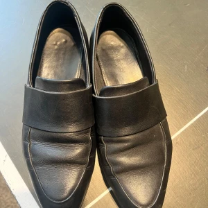 Svarta loafers från Vagabond - Snygga svarta loafers från Vagabond med stilren design och bred rem över foten. Skorna har spetsig tå och är tillverkade i mjukt skinn som ger en exklusiv känsla. Perfekta för dig som gillar klassiska och minimalistiska skor med en modern twist.