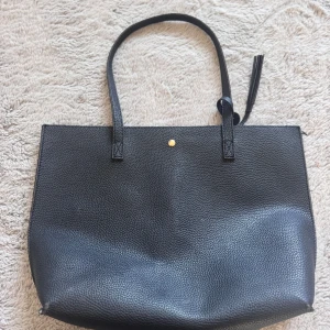 Svart axelväska i skinn  - Stilren svart axelväska i skinn med en enkel, fyrkantig form och en liten guldfärgad knapp framtill. Väskan har två långa axelremmar och en dekorativ tofs. Insidan är rymlig och perfekt för att få plats med allt du behöver. Köpt från shein.