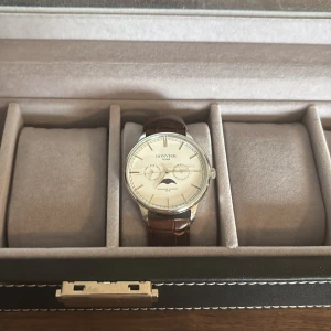 Bonvier Roma klocka med brun läderrem - Stilren Bonvier Roma klocka med rund silverfärgad boett och ljus urtavla. Klockan har en klassisk brun läderrem med krokodilpräglat mönster och detaljer som datum- och veckovisning samt månfas. Nypris 2100kr 