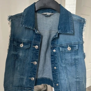 Blå jeansväst med fransade kanter - Säljer en blå jeansväst från Lindex med fransade kanter vid ärmhålen och klassisk krage. Västen har två bröstfickor med silverfärgade knappar och en rak, loose passform. Perfekt att styla över en hoodie eller t-shirt för en cool look. Främst för Barn 