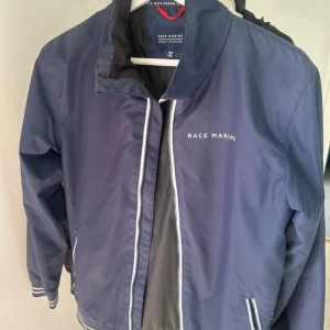 Blå vindjacka Race Marine 158/164 - Snygg marinblå vindjacka från Race Marine med vit logga på bröstet. Jackan har dragkedja framtill, ribbade muddar med vita ränder och två sidofickor med dragkedja. Perfekt för blåsiga dagar och har en sportig look som passar till jeans eller joggers.