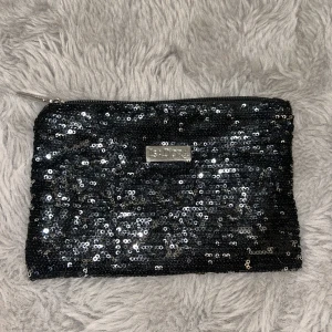 Svart glittrig neccesär från Isadora - Svart neccesär från Isadora med glittriga paljetter över hela ytan och silverfärgad dragkedja. Perfekt storlek för mobilen och småsaker. Neccesären har en metallbricka med Isadora-loggan framtill och insidan är fodrad i svart tyg.