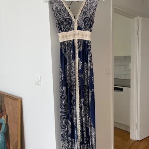 Marinblå långklänning - Säljer en marinblå vacker långklänning med vitt och blått mönster. Klänningen har slits, spetsdetaljer och knytband i ryggen. Midjan är markerad med bred spets och tyget är mjukt och följsamt