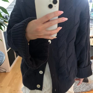 Mörkblå kabelstickad kofta med knappar - Säljer den populära mörkblå kabelstickade kofta från Gina. Koftan har rund hals och långa ärmar med breda ribbade muddar. Perfekt för att hålla sig varm under kyliga dagar