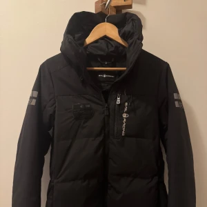 Svart dunjacka från Sail Racing M - Säljer en svart dunjacka från Sail Racing med huva och snygga logodetaljer på bröstet och ärmarna. Jackan har dragkedja framtill och är fodrad med Allied Feather + Down, 500 fill power. Perfekt för kalla dagar och har en clean, sportig look.