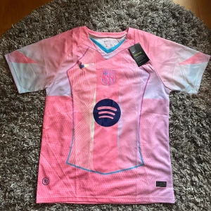 Barcelona rosa fotbollströja Nike - Snygg Barcelona fotbollströja från Nike i rosa och ljusblå toner. Tröjan har korta ärmar, V-ringad hals och coola grafiska linjer på framsidan. Spotify-logga och klubbmärke på bröstet. Perfekt nu i sommar. Storlek L sitter som M