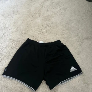 Svarta Adidas träningsshorts - Svarta träningsshorts från Adidas med vit logga på vänster ben och vita detaljer längs bensluten. Modellen har elastisk midja och är gjorda i lätt polyester som andas. Perfekta för sport eller gym och har en klassisk loose passform.